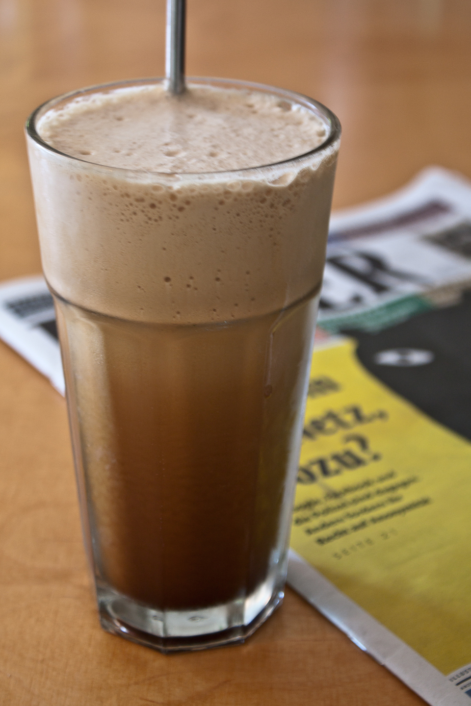 A tall glass of Greek Frappé, topped with a thick layer of frothy foam, with ice cubes and a straw, on a sunny outdoor table.
