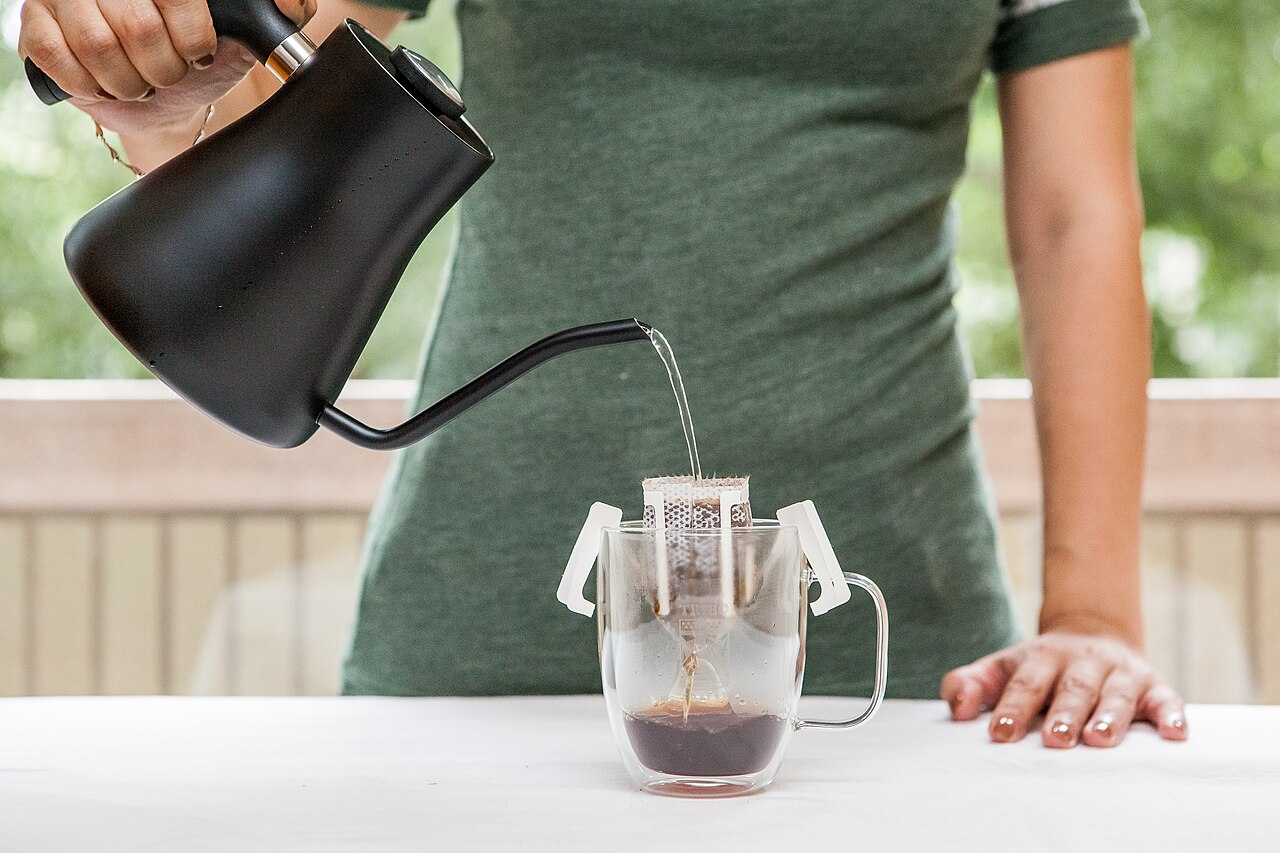 A close-up shot of a hand pouring hot water from a simple kettle onto ground coffee in a ceramic pour-over cone, with steam rising from the wet grounds.